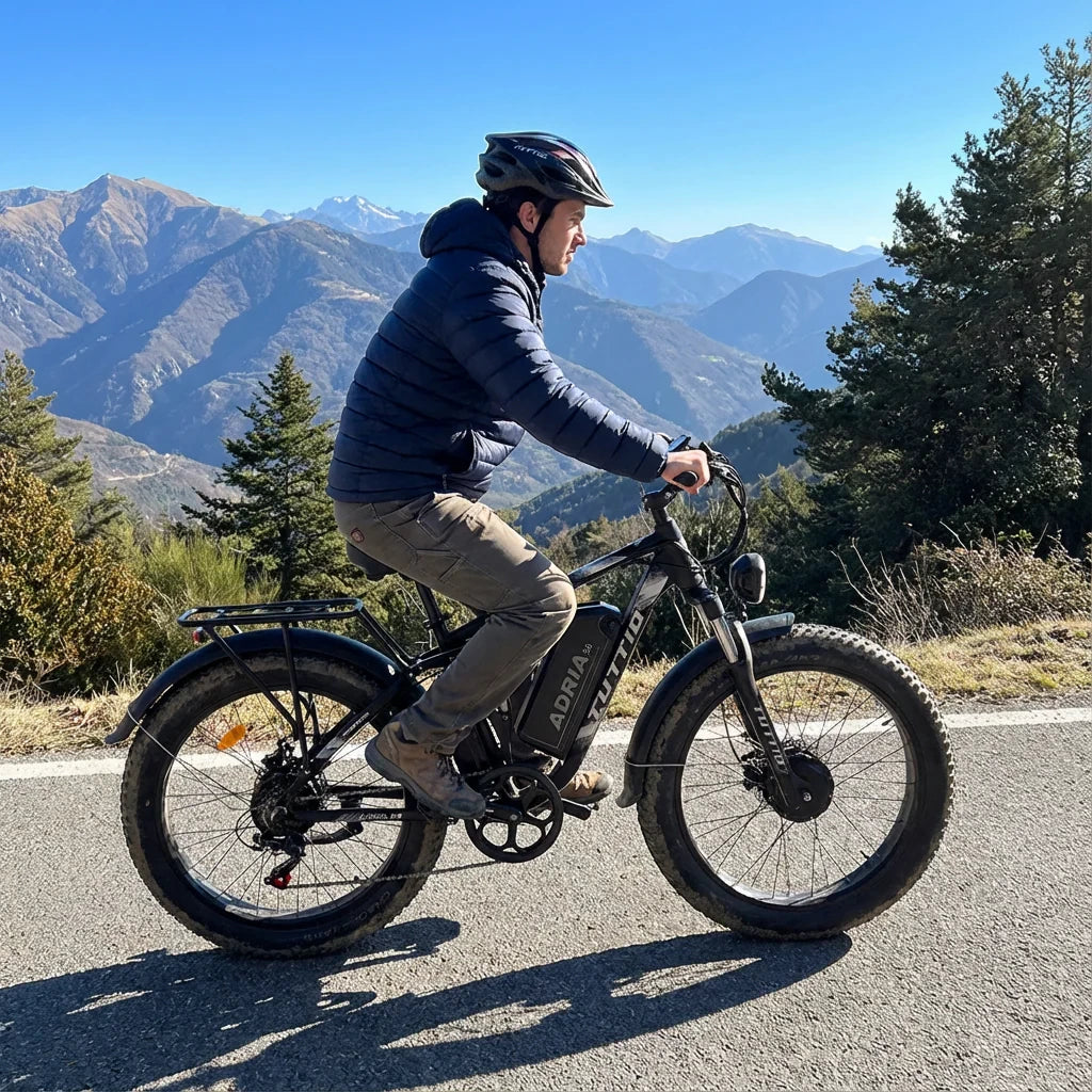 A man riding the Tuttio Adria26 fat tire electric bike on a paved scenic road with a beautiful mountain range in the background, highlighting the e-bike's versatility for smooth asphalt cruising.