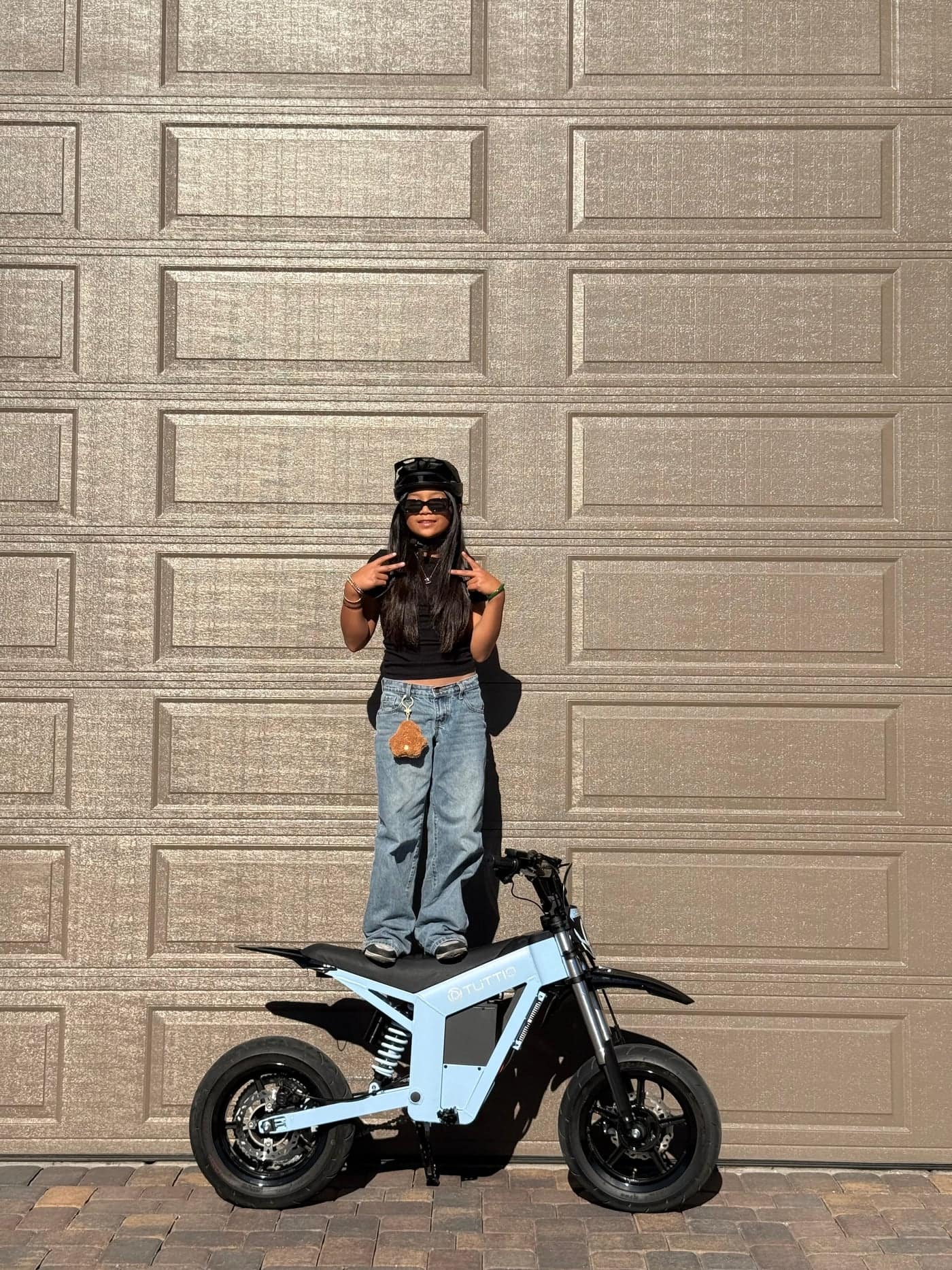 A sunny outdoor lifestyle shot featuring a stylish rider posing playfully by standing on the seat of the light blue Tuttio Soleil01 GB electric dirt bike in front of a garage door.