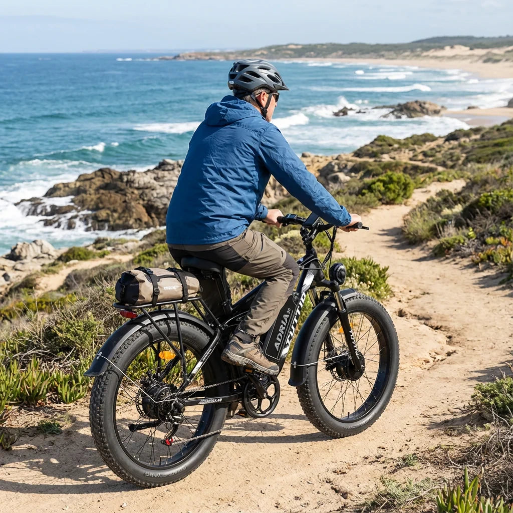 Helmeted rider on the Tuttio Adria26 riding along a coastal dirt trail, with the ocean in the background, black frame, black rims, fat tires, rear rack bag, and rear side riding posture visible