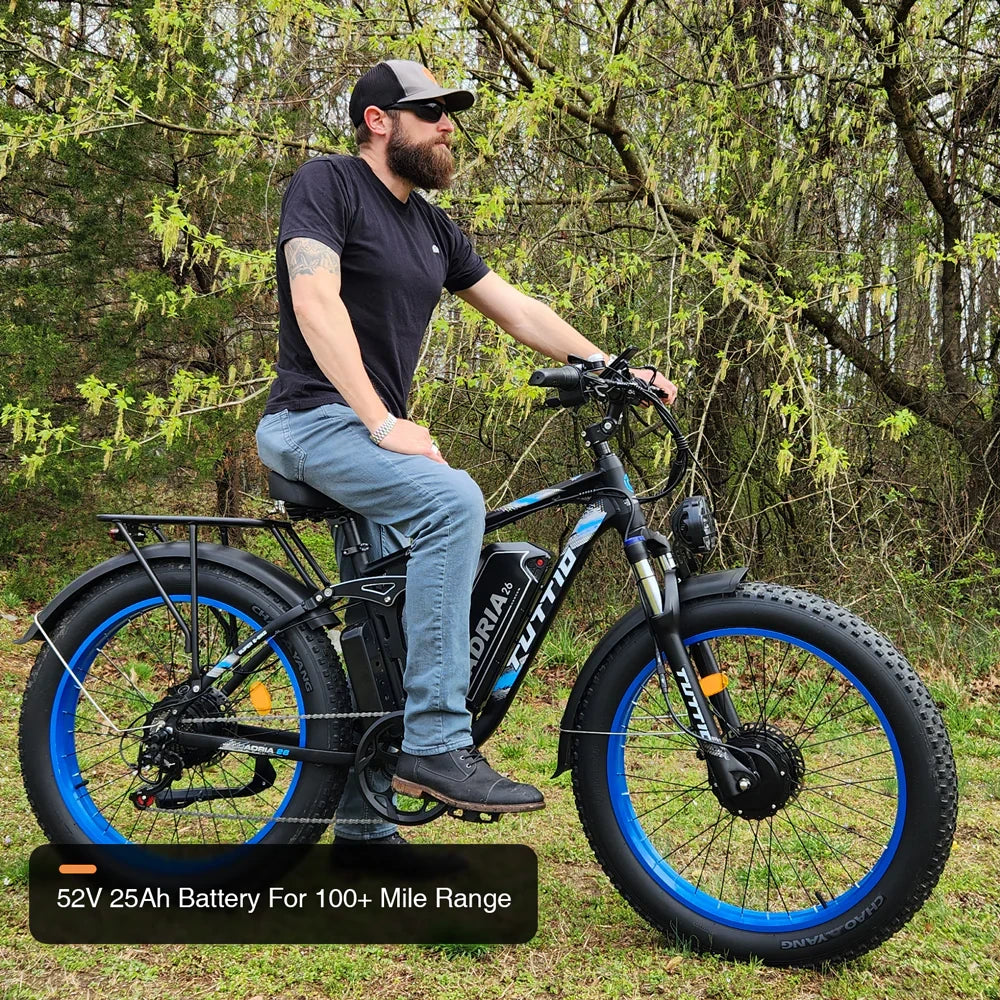 A bearded man sitting on the Tuttio Adria26 long-range e-bike with blue rims in a wooded area, featuring an overlay graphic that highlights its high-capacity 52V 25Ah battery capable of over 100 miles per charge.