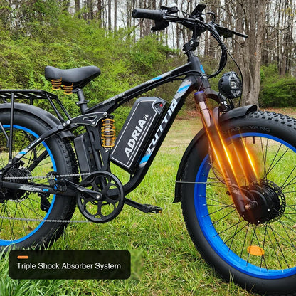 Close-up of the Tuttio Adria26 electric bike with blue rims parked on grass, featuring graphic overlays that highlight its triple shock absorber system including the front fork, rear frame, and seat springs.