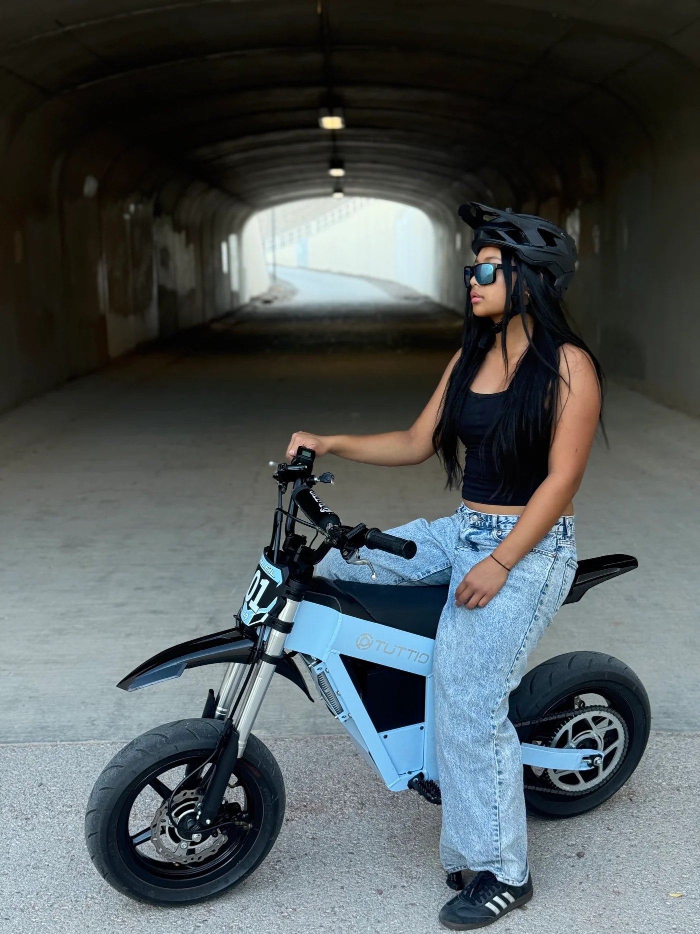 A rider in a helmet and sunglasses sitting on a light blue TUTTIO Soleil 01 GB electric mini dirt bike inside a concrete tunnel.