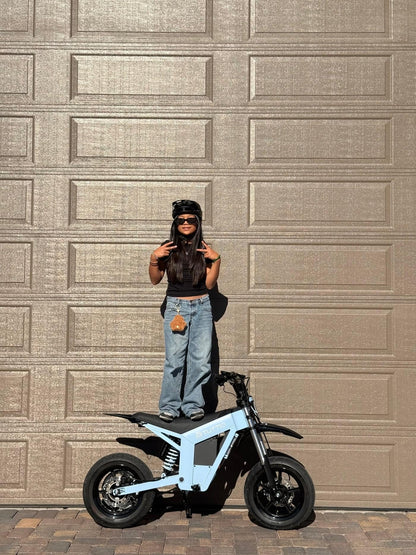 A sunny outdoor lifestyle shot featuring a stylish rider posing playfully by standing on the seat of the light blue Tuttio Soleil01 GB electric dirt bike in front of a garage door.