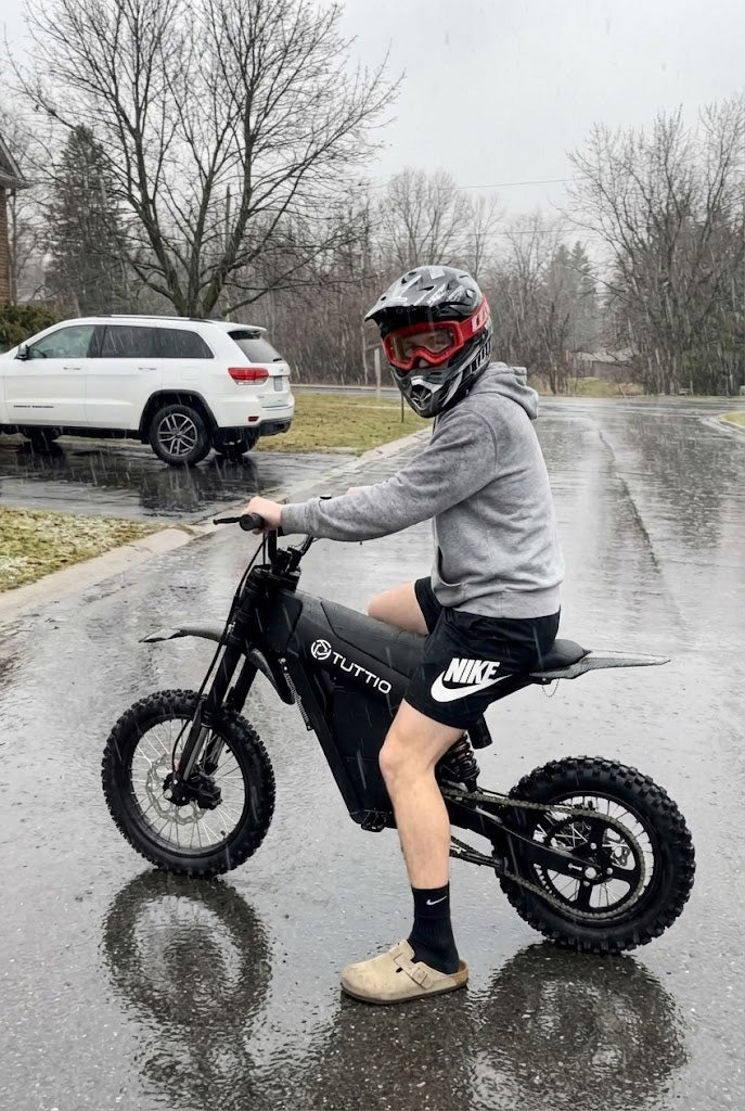 Customer photo of a rider sitting on a black Tuttio Soleil01 SE on a wet residential road, wearing a full-face helmet with red goggles, gray hoodie, black shorts, and showing the black frame, Tuttio logo, knobby off-road tires, road reflection, white SUV, and leafless trees in the background