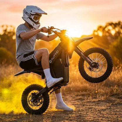 Rider wearing a white full-face helmet performing a wheelie on a black Tuttio Soleil01 SE electric dirt bike at sunset, showing the Tuttio frame logo, raised front wheel, knobby off-road tires, dirt trail, golden backlight, and dust behind the rear tire